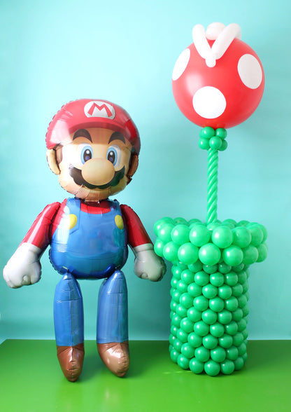 A Mario AirWalker foil balloon stands beside a balloon Piranha Plant in a green pipe against a blue backdrop.