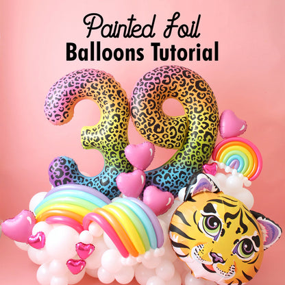 A vibrant balloon arrangement featuring custom spray-painted foil balloons shaped as "39" with a colorful leopard print design. The display is accented with rainbow balloon arches, pink heart-shaped balloons, and a tiger face foil balloon, all set against a soft pink background. This tutorial teaches how to transform standard foil balloons into unique, eye-catching designs using spray paint techniques.