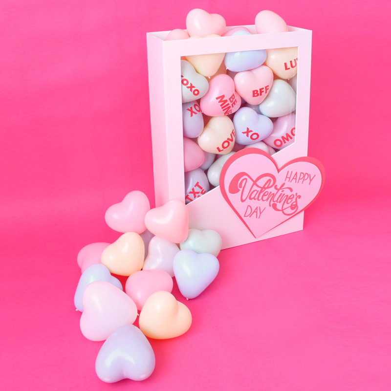 DIY Conversation Hearts Box balloon decor featuring a white Valentine’s display box overflowing with pastel heart balloons, styled on a bright pink background. The conversation hearts balloons include phrases like XOXO, LOVE, and BFF, creating a playful Valentine’s Day and Galentine’s balloon decor idea for party displays and themed event styling.