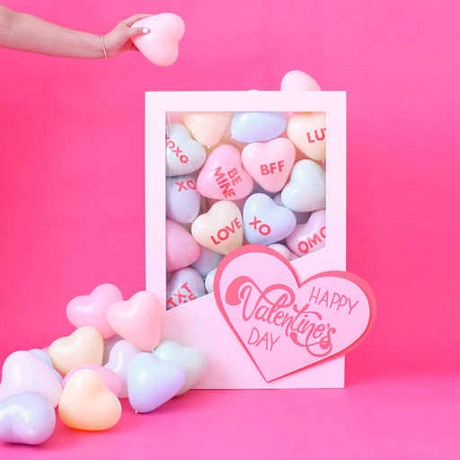 DIY Conversation Hearts Box balloon decor featuring a white display box filled with pastel heart balloons printed with phrases like XOXO, LOVE, BFF, and BE MINE, styled on a pink background for Valentine’s Day decor. This conversation hearts box is a playful balloon decoration idea for Valentine’s parties, Galentine’s Day celebrations, and themed event displays.