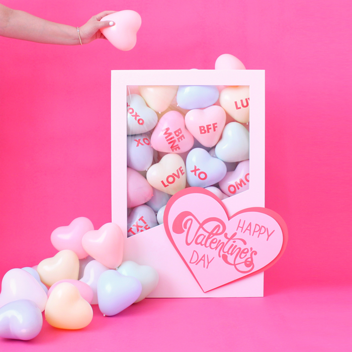 DIY Conversation Hearts Box balloon decor featuring a white display box filled with pastel heart balloons printed with phrases like XOXO, LOVE, BFF, and BE MINE, styled on a pink background for Valentine’s Day decor. This conversation hearts box is a playful balloon decoration idea for Valentine’s parties, Galentine’s Day celebrations, and themed event displays.