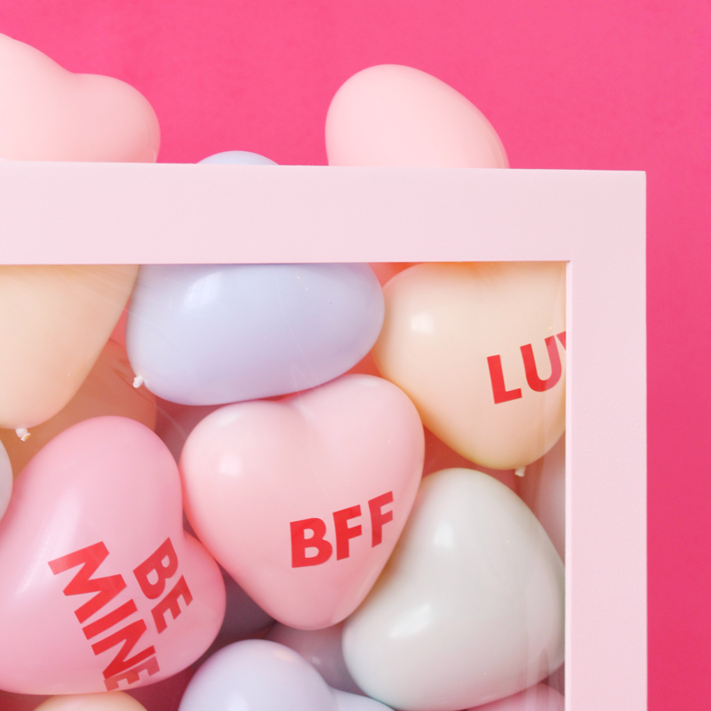 Close-up of a DIY Conversation Hearts Box showing pastel heart balloons printed with phrases like BFF, BE MINE, and LUV, framed inside a white display box on a pink background. This detail highlights the conversation hearts balloon decor used for Valentine’s Day and Galentine’s party displays and themed event decor.